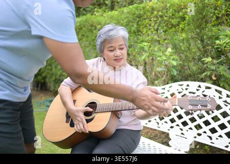 Senior woman sitting on a bench in the garden playing an acoustic guitar while a friend teaching. Concept of music learning and creative leisure in retirement || Model released Stock Photo