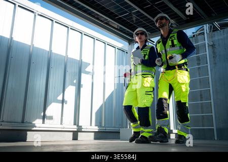 Male and female engineers inspecting photovoltaic systems beneath solar panel structures on a rooftop, representing professional renewable energy management || Model released Stock Photo