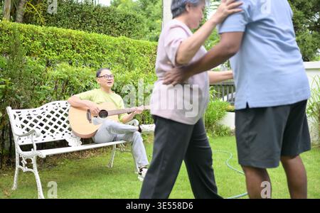Group of asian seniors singing and dancing while their friend plays guitar in a garden, enjoying friendship and outdoor leisure activity || Model released Stock Photo