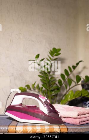 Modern pink-and-white iron rests on a checkered ironing board. Beside it: neatly folded pink towel and green houseplant against a light wall. Perfect Stock Photo
