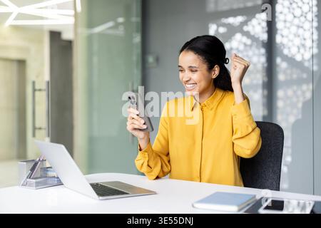 Happy young woman sitting at a desk in an office, celebrating an achievement or good news while looking at her smartphone and raising a fist in triumph Stock Photo