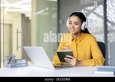 Young woman wearing headphones and a yellow shirt, smiling and writing notes in a small book while attending an online meeting or webinar using a laptop at a modern office desk Stock Photo