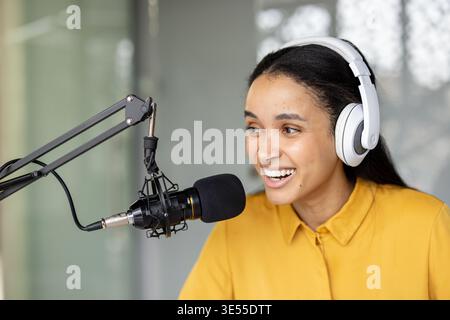 Young woman wearing headphones and speaking into a microphone, hosting a live podcast from her home studio, smiling while creating engaging audio content and streaming online Stock Photo