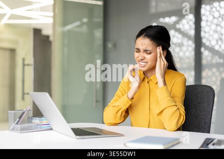 Young woman working on laptop in a modern office, feeling severe headache and pain, touching her temples with a pained expression due to stress and burnout Stock Photo