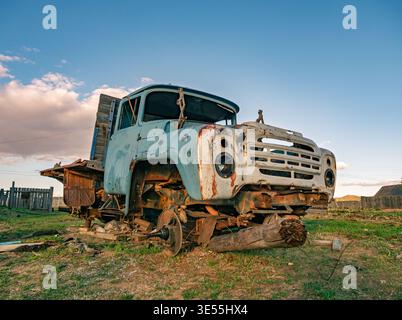 Old blue truck sits in field with wood logs under its front near abandoned wooden house and fence during late afternoon light on a clear day Stock Photo