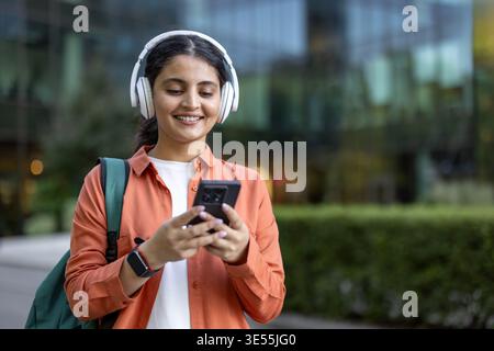 Young woman student listening to music or podcast through white headphones, smiling while looking at her smartphone and walking outdoors on a college campus with a backpack Stock Photo