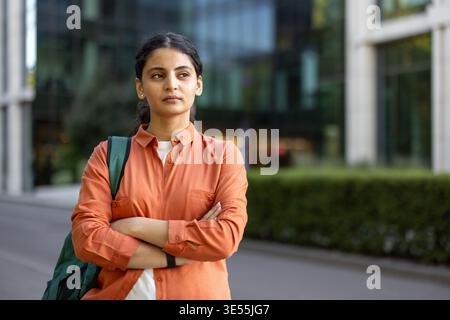 Young businesswoman with a backpack standing confidently with folded arms outside a modern university building, symbolizing ambition and future aspirations Stock Photo