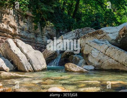 Flowing mountain river with clear water winding through a sunlit rocky gorge, surrounded by lush green trees and ancient stone formations Stock Photo