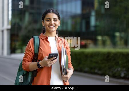 Young indian female student smiling, carrying a backpack and holding books and a smartphone, walking confidently on a university campus, representing education, success, and aspirations Stock Photo