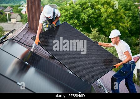 Workers building solar panel system on roof of house. Two men installers in helmets carrying photovoltaic solar module outdoors. Alternative, green and renewable energy generation concept. Stock Photo