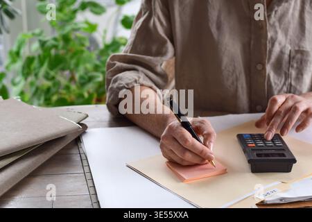 Woman writing on sticky notes notepad while using calculator at desk with 2025 taxes file folder. Home office scene focused on financial planning, bud Stock Photo