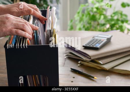 Person hands sorting through hanging file folders on desk with documents, calculator, and pen in home setting. Organized workspace highlighting paperw Stock Photo