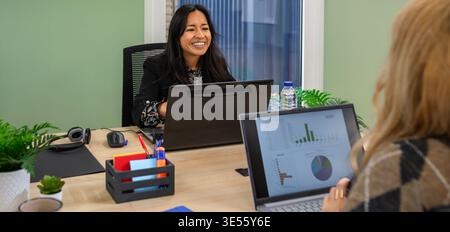 Portrait of smiling hispanic businesswoman talking with female colleague in a modern office space Stock Photo