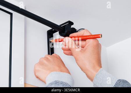 a man marks the drilling point on a white wall with a red pencil while holding a black metal bracket for a curtain rod in place near the corner of the Stock Photo