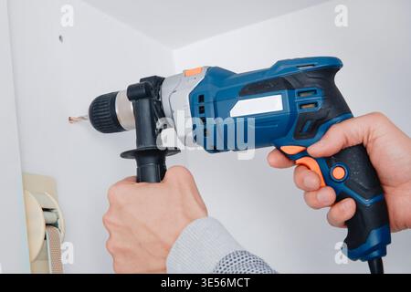 close view of a man using an electric drill to make a hole in a white wall inside a room corner, holding the tool firmly with both hands Stock Photo