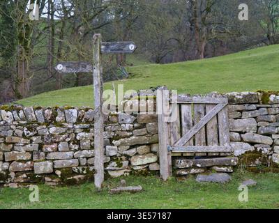 Yorkshire, UK - Mar 14 2026: Rustic waymarker beside a weathered wooden gate set in a dry stone wall in Yorkshire UK, with green pasture and trees creating a quiet rural scene Stock Photo