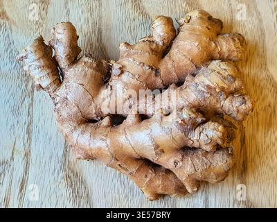 A fresh, unpeeled ginger rhizome (Zingiber officinale) resting on a light-colored wooden surface. The root displays its characteristic rough, tan to Stock Photo