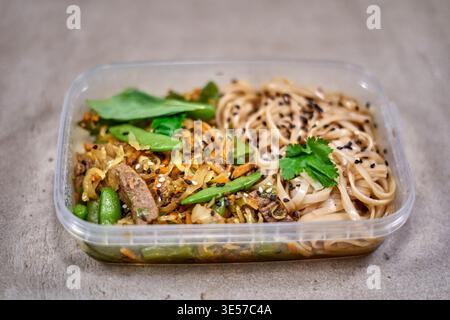 A transparent plastic rectangular meal prep container filled with an Asian-inspired stir-fry dish sits on a gray textured surface. The container is Stock Photo