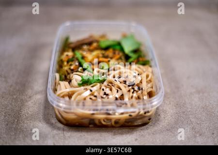 A close-up photograph of a rectangular transparent plastic takeaway container filled with an Asian-inspired noodle dish, placed on a textured gray Stock Photo