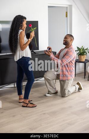 Diverse couple kneeling, man holding open ring box while woman holding red rose in living room Stock Photo