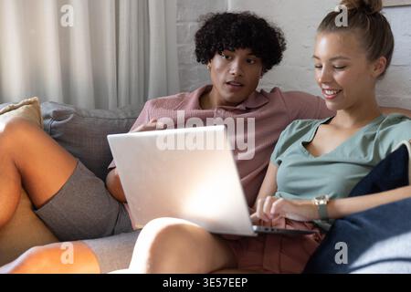 Couple sitting closely on gray sofa at home, sharing silver laptop and wearing wristwatch Stock Photo