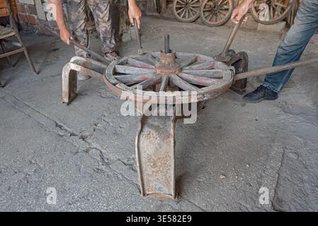 Traditional master blacksmith and wheelwright repairing a vintage wooden horse carriage wheel in a rustic workshop in Kula, Manisa Province, Turkey. Stock Photo