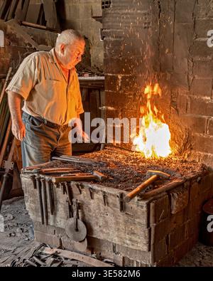 Veteran blacksmith heating metal rod in a blazing forge fire. Kula, Turkey - May 24, 2022: An elderly male artisan monitors the glowing iron within a Stock Photo
