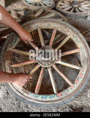 Detail shot showing hands working on a vintage, hand-painted wooden wagon wheel in a restoration workshop. The wheel features colorful patterns. Stock Photo