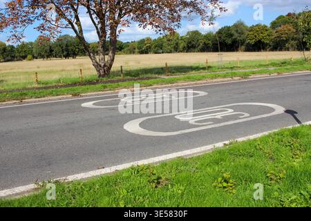 Two speed limit markings are painted on an asphalt road in a rural setting of English countryside, surrounded by green grass, a brown-leaved tree. Stock Photo