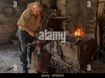 Master blacksmith hammering glowing hot metal on a traditional anvil. Kula, Turkey - May 24, 2022: An elderly craftsman strikes the iron in a historic Stock Photo
