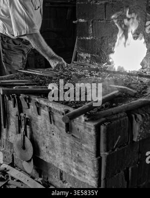 Blacksmith forging hot metal with sparks in a rustic forge. Medium shot of the metalworking process with hammers and tools on a stone anvil block. Stock Photo