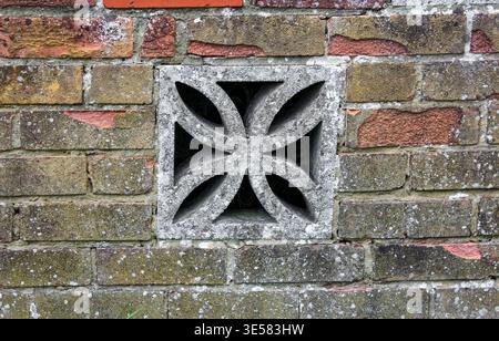An weathered, grey concrete breeze block with a floral geometric cutout is integrated into an old, multi-toned brick wall with moss and lichen. Stock Photo