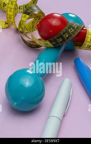 Blue and red dumbbells with a yellow measuring tape and pens on a purple background, representing a fitness plan and weight loss goals Stock Photo