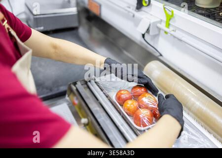A grocery store assistant using plastic wrap to neatly package fresh tomatoes. The image shows careful handling and presentation of produce in a fruit Stock Photo