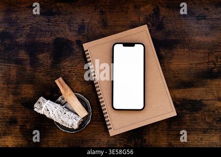 Smartphone with blank screen on notebook with sage and palo santo on wooden table, minimal workspace flat lay with natural elements. Top view Stock Photo