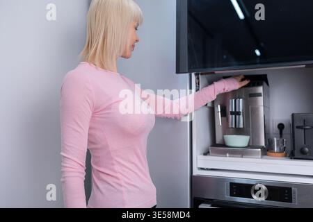 Coffee preparation. Adult blonde woman operating espresso machine holding empty glass cup in bright kitchen interior with focus on routine Stock Photo