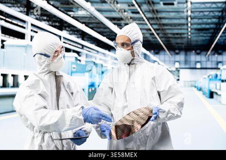Multiethnic men processing silicon wafers in automated manufacturing plant. Coworkers wearing bunny suits inspecting delicate semiconductor substrates hardware during quality control process Stock Photo