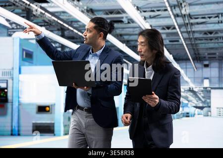 Multiracial businessmen in microchip facility brainstorming, overseeing hardware production metrics. Multiethnic teamworking colleagues using devices to monitor manufacturing gear bottlenecks Stock Photo