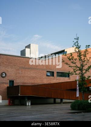 A renovated red brick industrial building featuring a striking diagonal rusty corten steel ramp, set against modern city skyscrapers under a blue sky. Stock Photo