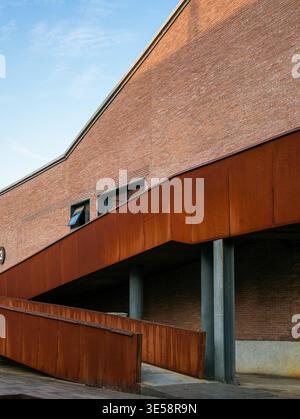 Close up of a diagonal rusty corten steel architectural ramp zigzagging across a renovated red brick building facade under a blue sky. The scene repre Stock Photo