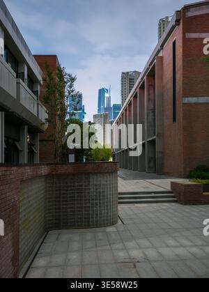 A curved red brick retaining wall along a paved pedestrian walkway flanked by low-rise buildings, leading toward a distant modern city skyline at twil Stock Photo