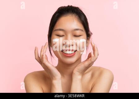 Young Woman Applying Skincare Cream on Her Face With a Smile in Front of a Pink Background Stock Photo