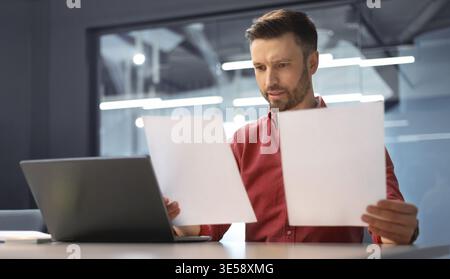 Man Reviews Documents While Using Laptop in Modern Office Setting During Daytime Hours Stock Photo
