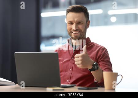 Man Giving Thumbs up While Sitting at Desk With Laptop and Coffee Cup in Office Setting Stock Photo