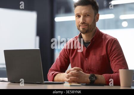 Young Man Sitting at Desk With Laptop and Notebook in Modern Office Setting Stock Photo