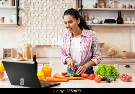 Woman Preparing Vegetables in Kitchen While Watching Cooking Video on Laptop Stock Photo