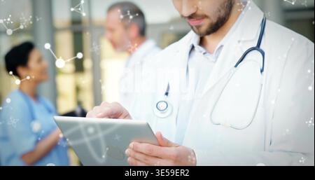 Using tablet, clinician in white coat, stethoscope reviewing data in corridor, nurse in blue scrubs Stock Photo