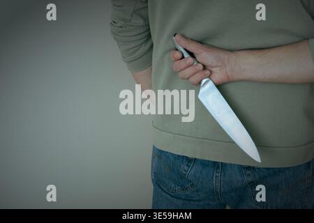 man holding a kitchen knife behind his back, suggesting danger, threat or hidden intent. Concept of violence, crime, secrecy or potential risk indoors Stock Photo