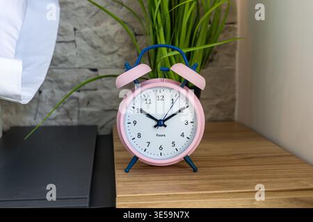 Close-up of a pink vintage alarm clock on wooden bedside table with green plant and modern bedroom background Stock Photo