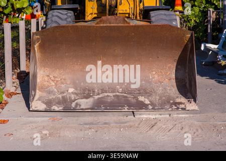 Heavy construction equipment attachment sits idle on a paved surface near a beach area. A medium close-up shot captures the curved steel blade. Stock Photo